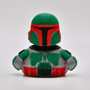May include: A green and silver rubber duck with a Boba Fett helmet. The duck has a red visor and a black mouth.