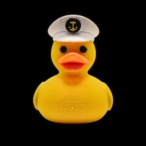 May include: A yellow rubber duck wearing a white sailor hat with a black anchor emblem. The duck has black eyes and an orange beak.