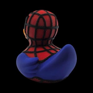 以下が含まれることがあります： スパイダーマン風のラバーダック。赤と黒のクモの巣模様のマスクと胴体、青色のアクセントが下半身に施されています。おもちゃは黒い背景に配置されています。