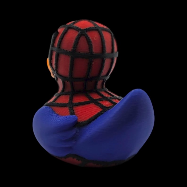 以下が含まれることがあります： スパイダーマン風のラバーダック。赤と黒のクモの巣模様のマスクと胴体、青色のアクセントが下半身に施されています。おもちゃは黒い背景に配置されています。