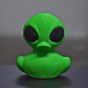 May include: A green alien-shaped figurine with large black eyes and a duck-like beak. The figurine is a bright, solid green color and has a textured surface. The alien has a rounded head and body, and is likely a decorative item.
