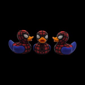以下が含まれることがあります： スパイダーマン風のラバーダック3羽。各アヒルは、体に赤と黒のウェブ模様、青い翼、黄色のくちばしが付いています。目は白で、背景は黒です。