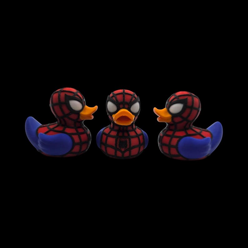 以下が含まれることがあります： スパイダーマン風のラバーダック3羽。各アヒルは、体に赤と黒のウェブ模様、青い翼、黄色のくちばしが付いています。目は白で、背景は黒です。