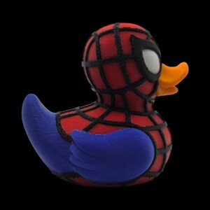 以下が含まれることがあります： スパイダーマン風のノベルティラバーダック。アヒルは、黒いクモの巣模様の赤い体、青い翼、オレンジ色のくちばし、白い目の黒いマスクが特徴です。おもちゃは黒い背景に配置されています。