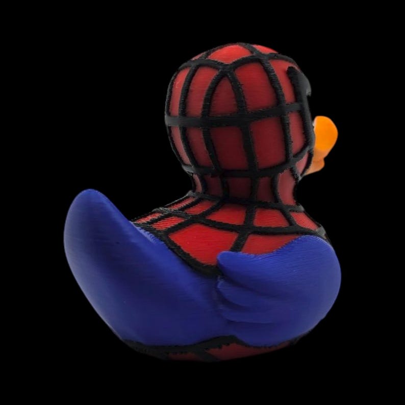 以下が含まれることがあります： スパイダーマンのデザインが施された、斬新なラバーダック。アヒルの頭と体は赤色で、黒いクモの巣模様、青い翼、オレンジ色のくちばしが付いています。おもちゃは黒い背景に配置されています。