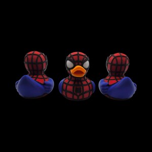 以下が含まれることがあります： スパイダーマン風のラバーダック3体。赤と黒のクモの巣模様のマスク、青いコスチューム、オレンジ色のくちばしが特徴です。黒い背景に配置され、スーパーヒーローをテーマにしたお風呂のおもちゃを展示しています。