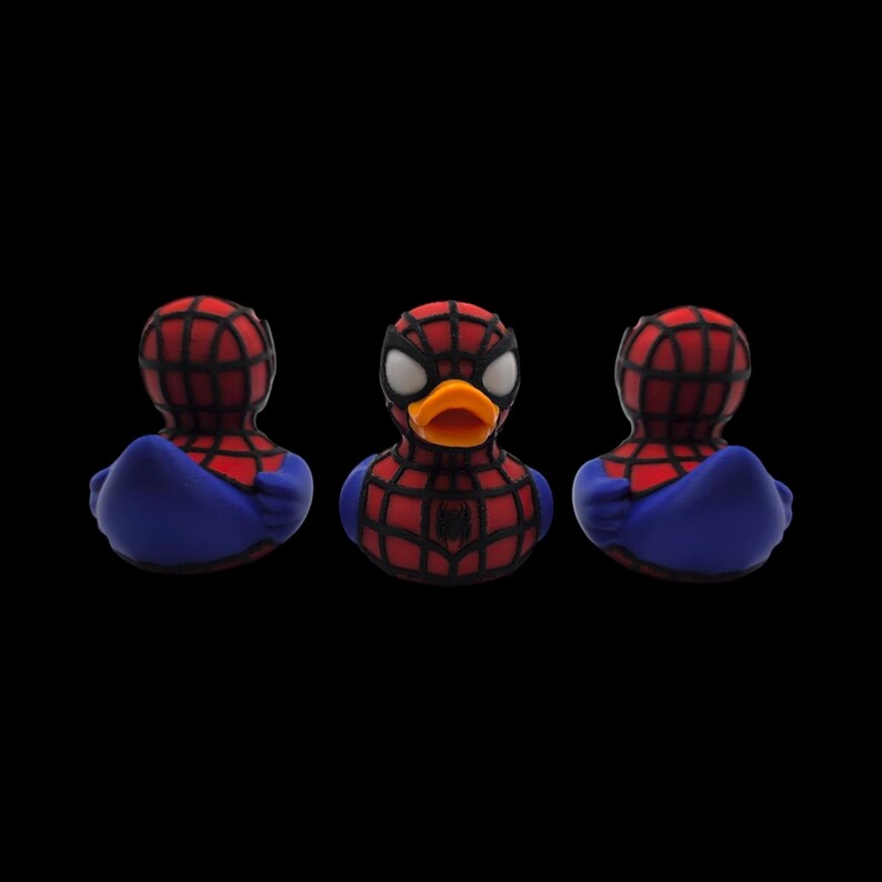 以下が含まれることがあります： スパイダーマン風のラバーダック3体。赤と黒のクモの巣模様のマスク、青いコスチューム、オレンジ色のくちばしが特徴です。黒い背景に配置され、スーパーヒーローをテーマにしたお風呂のおもちゃを展示しています。