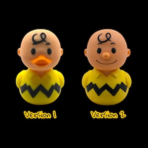 May include: Two novelty figurines, one a duck-like character and the other a human-like character, both in yellow shirts with black zigzags. The duck version has an orange beak. The human version has a smiling face. The text "Version 1" and "Version 2" are below each figurine.