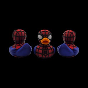 以下が含まれることがあります： スパイダーマンのデザインのゴム製アヒルが3羽。アヒルの頭と胸には赤と黒のクモの巣模様があり、青い翼が付いています。中央のアヒルは白い目とオレンジ色のくちばしをしています。