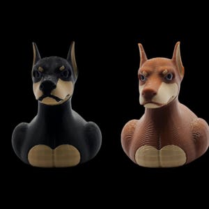 May include: Two dog busts, one black and one brown, with tan markings on their faces and chests. The dogs have pointed ears and a stern expression, set against a black background. The busts are likely decorative items.