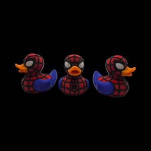 以下が含まれることがあります： スパイダーマンを模した3つのラバーダック。アヒルは赤色で黒いクモの巣模様、青い翼、白い目があります。オレンジ色のくちばしがあり、黒い背景に配置されています。