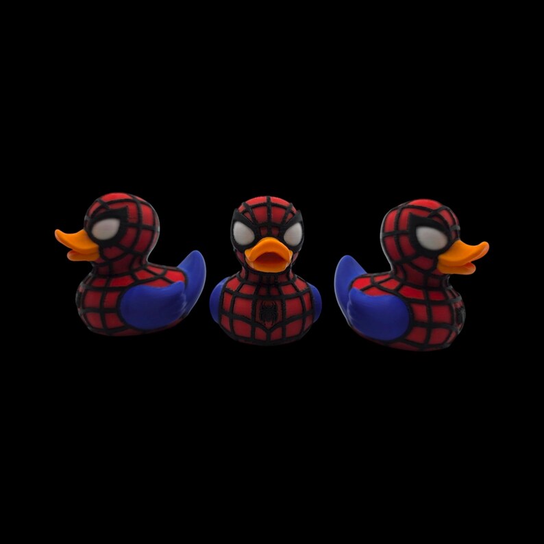 以下が含まれることがあります： スパイダーマンを模した3つのラバーダック。アヒルは赤色で黒いクモの巣模様、青い翼、白い目があります。オレンジ色のくちばしがあり、黒い背景に配置されています。