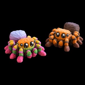 May include: Two colorful, cartoon-style spider figurines. One spider has a rainbow-colored body and legs, and a light purple head. The other spider is orange and brown. Both have large, round eyes and are set against a black background.
