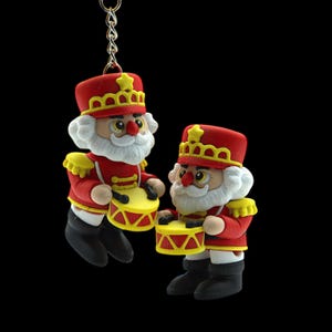 Peut inclure: Deux porte-clés sur le thème de Noël représentant des soldats jouets batteurs. Chaque figurine porte un uniforme rouge avec des détails jaunes, un chapeau rouge avec une étoile dorée et des bottes noires. Ils ont des barbes blanches et tiennent des tambours jaunes avec des détails rouges. Le fond est noir.