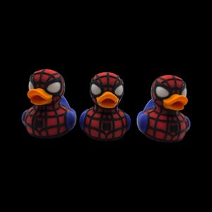 以下が含まれることがあります： スパイダーマン風のラバーダックのおもちゃが3つ。各アヒルは赤と黒のクモの巣模様、白い目、オレンジ色のくちばしを持っています。アヒルは青い翼と胸にスパイダーマンのロゴが付いています。背景は黒です。