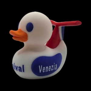 May include: A white duck-shaped figurine with blue eyes and a bright orange beak. The duck has a red and white tail fin and a blue oval with the word "Venezia" printed on it. The word "ival" is also printed on the duck.