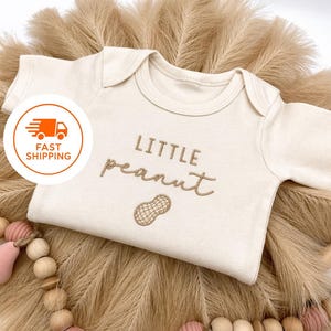 May include: Cream-colored baby onesie with the embroidered text "LITTLE peanut" and a peanut graphic. The onesie is displayed on a bed of dried, tan-colored grass and surrounded by a wooden bead garland with pink accents.