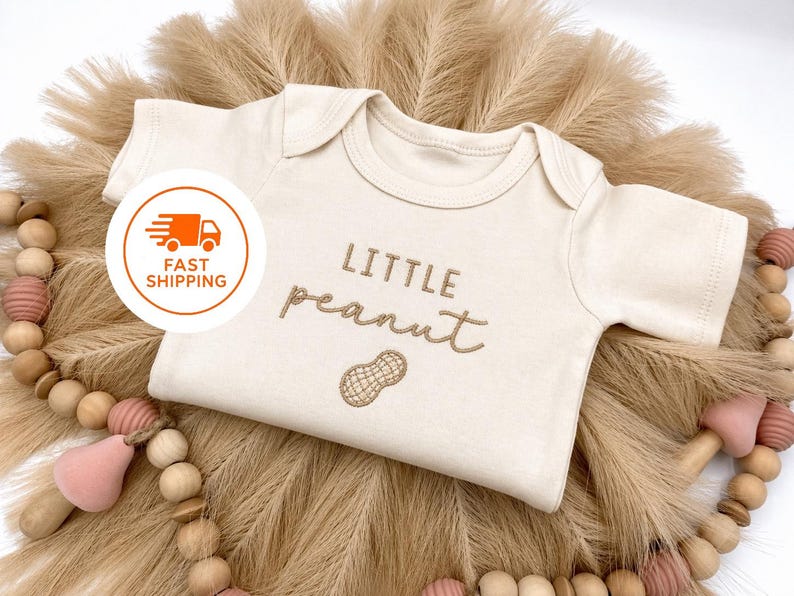 May include: Cream-colored baby onesie with the embroidered text "LITTLE peanut" and a peanut graphic. The onesie is displayed on a bed of dried, tan-colored grass and surrounded by a wooden bead garland with pink accents.