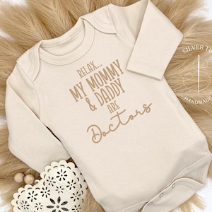 May include: A beige long-sleeved bodysuit with the text "Relax... My Mommy & Daddy Are Doctors" printed on the front.
