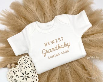 May include: White bodysuit with short sleeves and the text "NEWEST Grandbaby COMING SOON" in brown lettering.