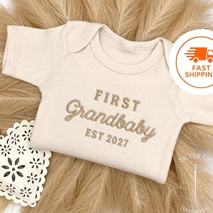 May include: Cream-colored baby onesie with the embroidered text "FIRST Grandbaby EST 2027". The onesie is displayed on a neutral background with decorative elements. A fast shipping icon is visible.