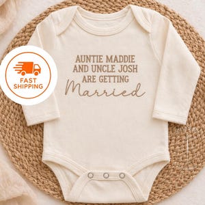 May include: Cream-colored baby onesie with long sleeves. The text "Auntie Maddie and Uncle Josh are getting Married" is printed in brown script. The onesie is placed on a woven mat.