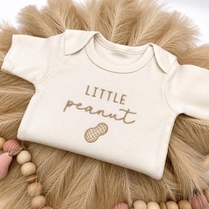 May include: Cream-colored baby onesie with the embroidered text "LITTLE peanut" and a peanut graphic. The onesie is displayed on a bed of dried, tan-colored decorative grass, with a wooden bead garland and pink accents.