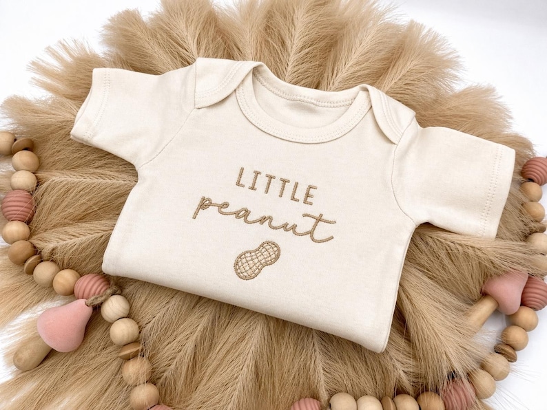 May include: Cream-colored baby onesie with the embroidered text "LITTLE peanut" and a peanut graphic. The onesie is displayed on a bed of dried, tan-colored decorative grass, with a wooden bead garland and pink accents.