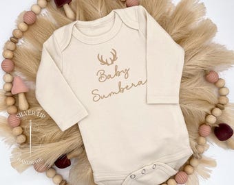 May include: A cream-colored long-sleeved baby onesie with a brown deer antler graphic and the text "Baby Sunbera".