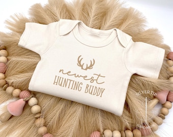 May include: A cream-colored baby onesie with the text "newest hunting buddy" and a brown deer antler graphic.