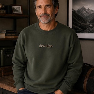 May include: An olive green crewneck sweatshirt featuring the word grandpa embroidered in a light tan script font across the center of the chest. The garment has long sleeves with ribbed cuffs and a ribbed neckline, constructed from a solid fabric.