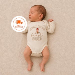 May include: A cream-colored long-sleeve baby bodysuit featuring an embroidered graphic of a red and white cowboy boot. The text reads, "ACTUALLY... THIS IS MY first RODEO" in brown lettering. The garment has a round neckline and snap closures.