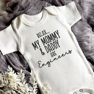 May include: White baby onesie with black text that reads "Relax... My Mommy & Daddy Are Engineers".