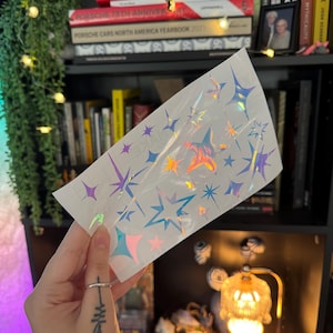 May include: A sheet of holographic star stickers in various sizes and shapes. The stickers reflect a rainbow of colors, including pink, purple, and blue. The stickers are on a white backing and are ready to peel and apply.