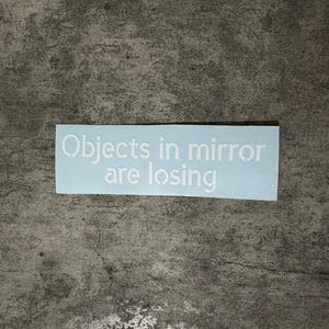 Puede incluir: Una pegatina rectangular azul claro con el texto "Objects in mirror are losing" en blanco. La pegatina está sobre una superficie gris texturizada.
