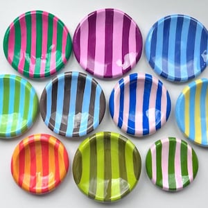 May include: Ten circular ceramic dishes arranged in a group, each featuring a distinct color combination of vertical stripes. The bowls display various palettes including green and pink, purple and pink, blue and light blue, and orange and yellow.