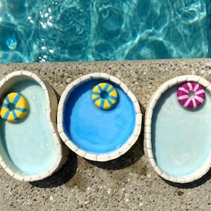 May include: Three ceramic dishes shaped like swimming pools with white tiled edges. Two dishes are oval and one is circular. The interiors are painted light blue. Each dish contains a small, floating ring-shaped toy in yellow and blue or pink.