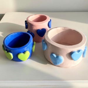 May include: Three ceramic planters in shades of blue and pink, decorated with heart-shaped accents. The blue planter has green hearts, while the pink planters feature blue hearts. The planters are arranged on a white surface.