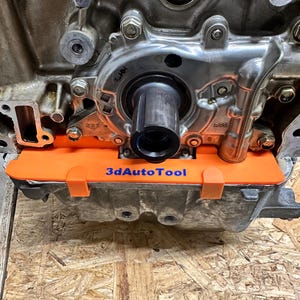 May include: An engine component with an orange 3dAutoTool part installed. The engine is silver with various bolts and a black cylindrical component. The orange part has the text "3dAutoTool" in blue.
