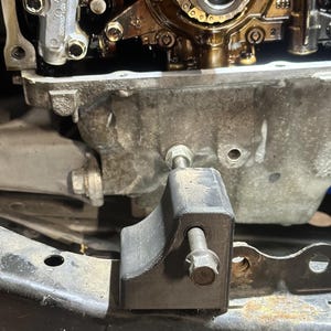 May include: A black 3D-printed plastic engine timing tool block with a textured surface, featuring a bolt inserted through the side. The tool is positioned against a metallic engine block component with visible gears, chains, and oil residue.