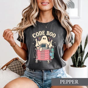 Puede incluir: Una camiseta gris carbón con un gráfico de un fantasma detrás de un carrito médico rosa con un monitor cardíaco, un estetoscopio y un tanque de oxígeno. El texto "CODE BOO" está impreso en una fuente amarilla clara sobre el fantasma.