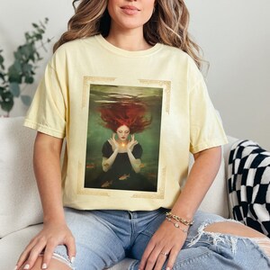 May include: A yellow t-shirt with a vintage-style illustration of a woman with red hair underwater. The illustration is framed with a gold border and features a few small gold fish.