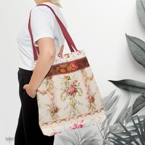 Può includere: Una borsa tote floreale con cinghie bordeaux. La borsa presenta uno sfondo color crema con un motivo ripetuto di rose rosa, foglie verdi e nastri blu. Una fascia marrone con accenti floreali e un bordo in pizzo adorna la parte superiore e inferiore.