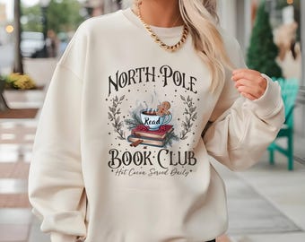 Könnte beinhalten: Cremefarbenes Sweatshirt mit einem Aufdruck "North Pole Book Club", der eine Tasse heiße Schokolade, einen Lebkuchenmann und Bücher zeigt. Der Text "Hot Cocoa Served Daily" ist ebenfalls enthalten. Das Sweatshirt hat einen lässigen Stil.