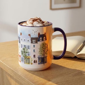 以下が含まれることがあります： パリの街並みが描かれたセラミックマグカップ。ホイップクリームとココアパウダーが入った飲み物が入っています。マグカップには青いハンドルとリムがあり、木製の表面に置かれています。