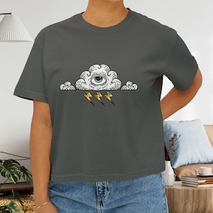 May include: A dark grey t-shirt featuring a graphic of a white cloud with a blue eye and three yellow lightning bolts.