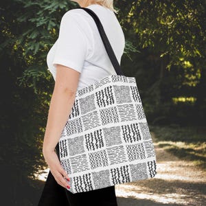 May include: A white tote bag with a black and white geometric pattern. The bag has a black shoulder strap and is carried by a person. The pattern consists of repeating shapes and lines, creating a visually interesting design.
