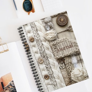 May include: A spiral-bound journal with a textured cover featuring a knitted design in shades of cream and gray. The cover includes wooden buttons and the word "Journal". The journal is open, revealing blank pages.