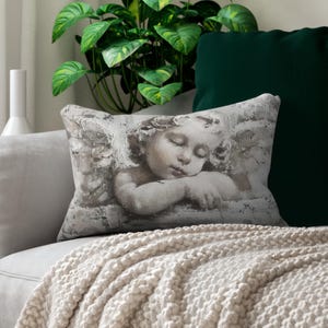 Puede incluir: Una almohada decorativa con un diseño gris y blanco que presenta un ángel querubín dormido con alas. La almohada está en un sofá blanco con una manta blanca.