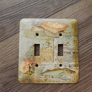 May include: A light switch cover with a vintage nautical theme. The cover features a sea turtle, a fish, and a seashell on a map-like background. The switch cover has two toggle switches.